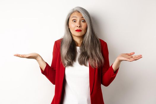 Confused asian senior woman shrugging, spread hands sideways and staring questioned at camera, dont know anything, standing over white background.