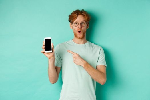 Check this out. Handsome redhead guy in glasses pointing finger at blank smartphone screen, showing online promotion, standing amazed over turquoise background.