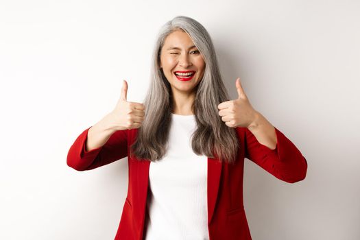 Cheerful asian mature woman winking, smiling pleased and showing thumbs-up in approval, like and agree, standing against white background.