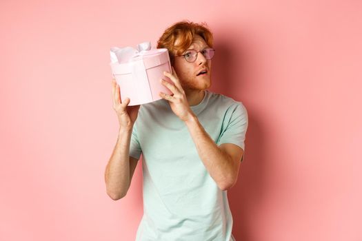 Valentines day and romance concept. Handsome redhead man shaking gift box and wonder what inside, trying to guess present, standing over pink background.