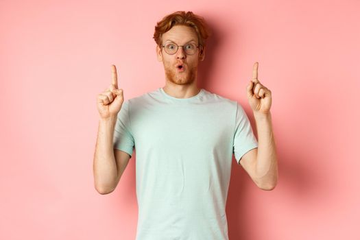 Impressed caucasian man with ginger hair, wearing glasses and t-shirt, saying wow and pointing fingers up at awesome deal, standing over pink background.