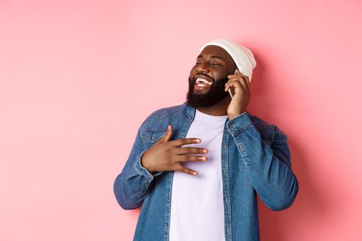 Happy african-american man talking on mobile phone, laughing and smiling, standing in beanie and denim shirt over pink background.