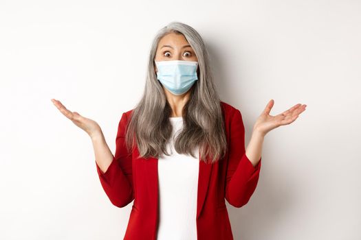 Covid-19 and business people concept. Asian businesswoman in face mask looking surprised, staring at camera in awe, standing over white background.