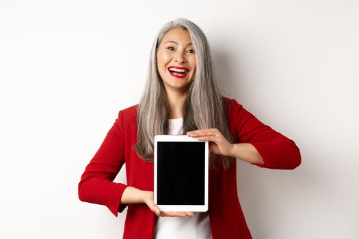 Business. Cheerful asian businesswoman in red blazer, showing digital tablet screen and smiling, introduce app or promotion, white background.