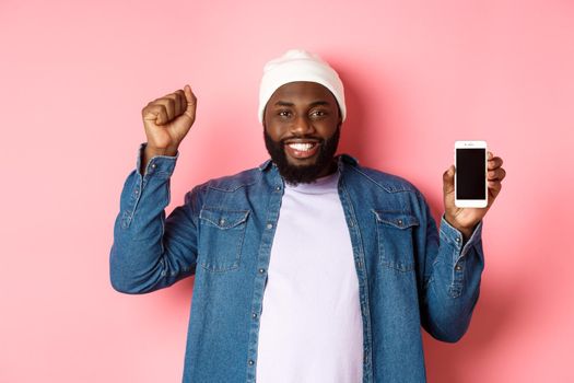 Online shopping and technology concept. Cheerful Black man rejoicing and showing mobile screen, raising hand up satisfied, triumphing while standing over pink background.