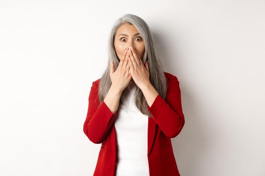 Shocked asian senior lady gasping, cover mouth from amazement, hear gossip, standing over white background.