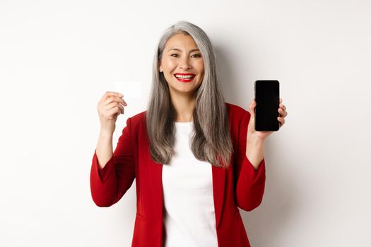 Senior asian businesswoman showing plastic credit card and blank smartphone screen, smiling at camera, white background.