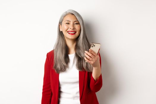 Asian female entrepreneur in red blazer using smartphone, smiling happy at camera, standing over white background.