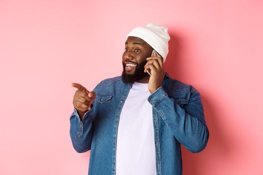 Handsome modern Black man talking on mobile phone, pointing left at person and smiling, standing over pink background.