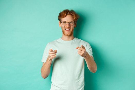 Handsome young man with ginger hair, wearing glasses and t-shirt, pointing finger at camera and smiling, choosing you, congratulating or praising, standing over mint background.