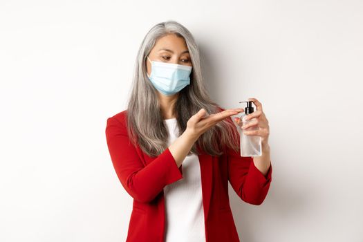 Covid, pandemic and business concept. Businesswoman in red blazer and face mask using hand sanitizer to clean and disinfect, standing over white background.