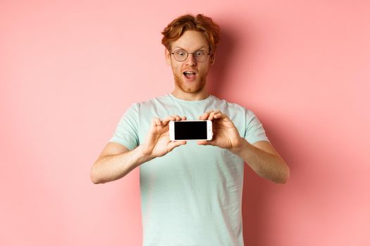 Amazed redhead man gasping and staring with awe phone, showing blank smartphone screen horizontally, standing over pink background.