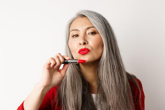 Beauty and makeup concept. Beautiful asian elderly woman pucker lips, showing red lipstick and looking sassy at camera, white background.