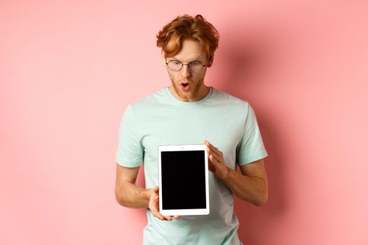 Impressed redhead man in glasses, showing blank digital tablet screen and looking in awe at display, saying wow, standing in t-shirt against pink background.