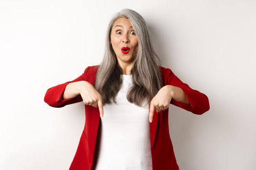 Portrait of happy asian lady in red blazer showing logo, pointing fingers down and smiling cheerful, check this out gesture, white background.