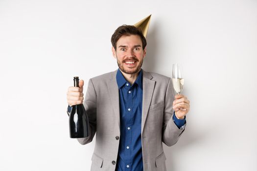 Celebration and holidays concept. Excited handsome birthday guy in party hat smiling, drinking champagne and having fun, white background.