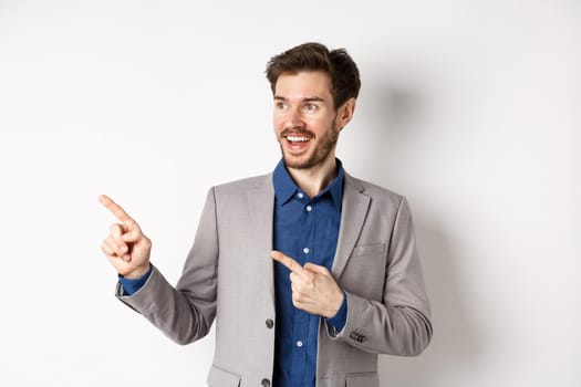 Excited bearded guy in festive suit pointing and looking left, smiling amazed, showing cool promo deal, standing on white background.