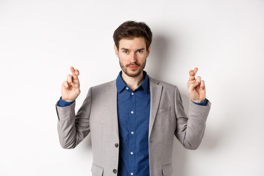 Hopeful man in suit cross fingers for good luck and looking confident in win, feeling determined, standing on white background.