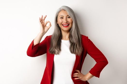 Asian professional businesswoman in red blazer, showing OK sign and smiling, approve and like something, standing against white background.