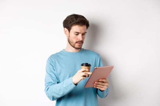 Man browsing online in digital tablet and drinking coffee from paper cup, standing over white background. Copy space