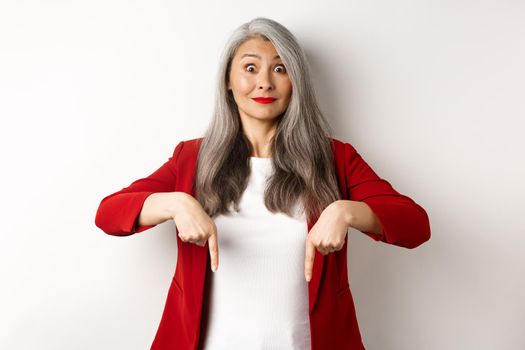 Beautiful asian senior woman in elegant red blazer, pointing fingers down and smiling excited, showing advertisement, white background.