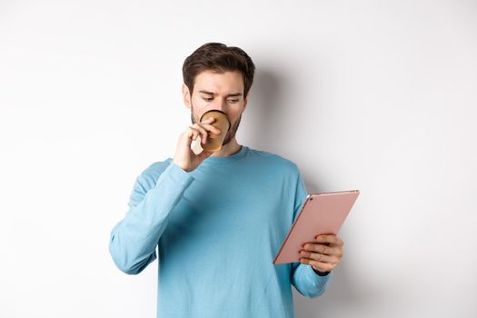 Handsome caucasian man drinking coffee and reading digital tablet screen, standing in blue sweatshirt over white background.