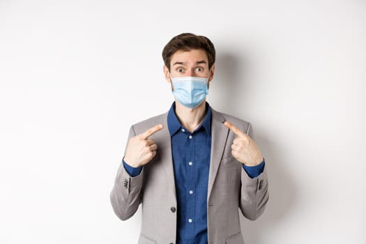 Covid-19, pandemic and business concept. Excited businessman in suit pointing at his medical mask and looking at camera, white background.