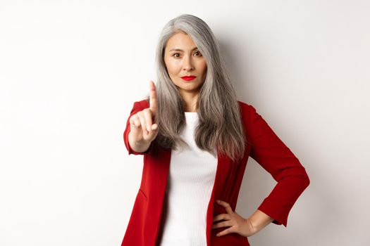 Serious and disappointed asian businesswoman showing stop gesture, shaking finger in disapproval, rejecting something bad, standing over white background.