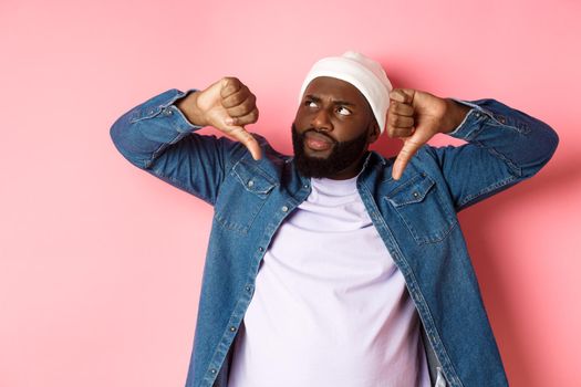 Disappointed Black man staring at upper left corner annoyed, showing thumbs-down, dislike something, standing over pink background.