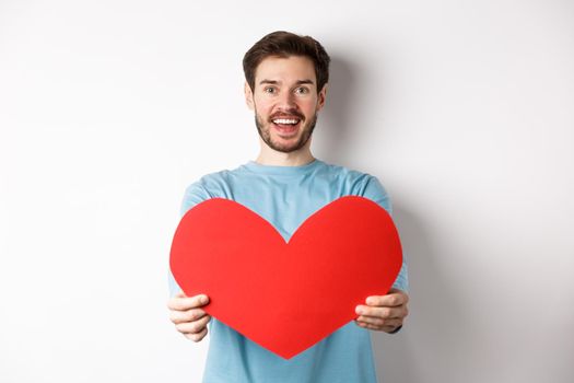 Relationship and people concept. Handsome man give valentines heart to you, smiling and saying I love you at camera, standing over white background.