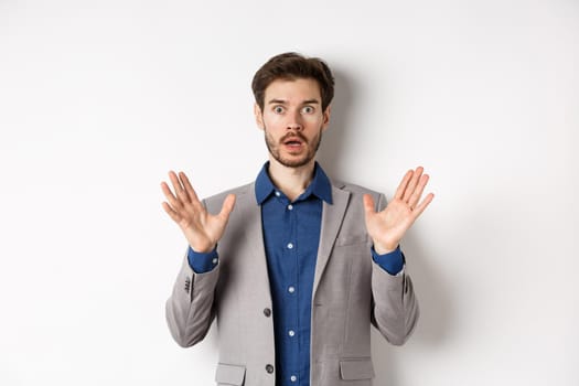 Surprised businessman in suit raising hands up and look excited, standing against white background.