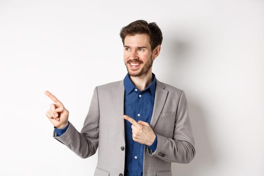 Successful businessman in grey suit pointing fingers left and looking at banner, smiling confident, showing advertisement, standing against white background.