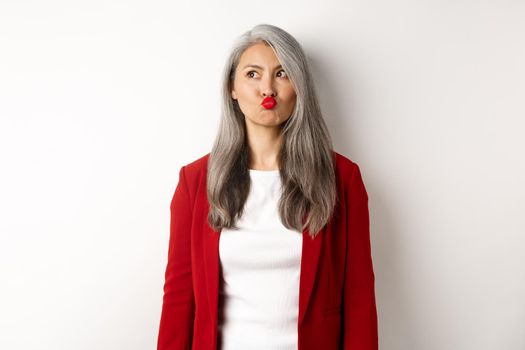 Indecisive asian businesswoman pucker lips and looking thoughtful at upper left corner, standing over white background.