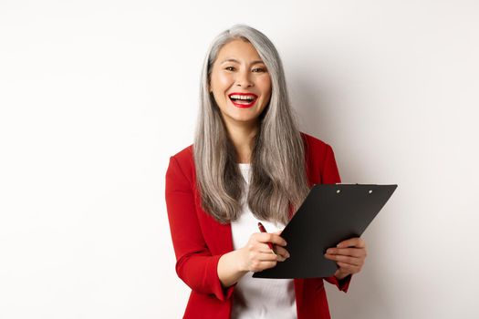 Happy asian office employee, woman in red blazer, working and laughing, holding clipboard with documents, standing over white background.