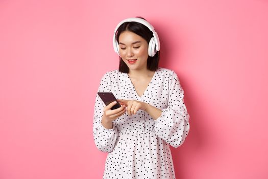 Beautiful asian woman texting message on smartphone, listening music in headphones, standing over pink background.