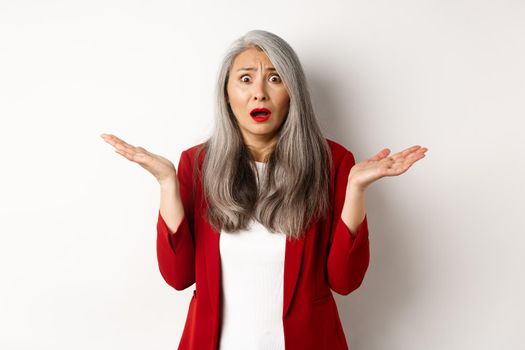 Business people. Confused and anxious asian senior office lady looking worried, spread hands sideways puzzled, standing over white background.