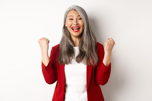 Lucky asian businesswoman achieve success, winning prize and celebrating, saying yes with fist pumps, standing happy against white background.