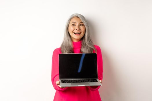 E-commerce concept. Happy beautiful asian senior woman with grey hair, looking aside and smiling, showing laptop black screen, standing over white background.