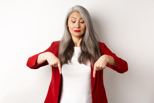 Intrigued asian elegant woman looking and pointing fingers down, showing logo, standing in red blazer over white background.