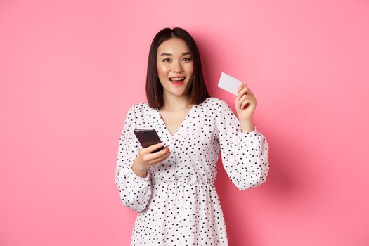 Attractive young asian woman order online, holding credit card and mobile phone, making internet purchase, standing happy over pink background.
