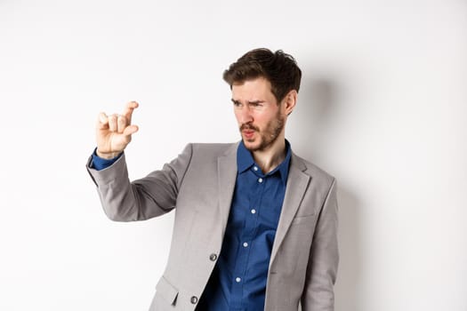 Businessman trying to see something small, showing little thing size with fingers, standing in suit on white background.