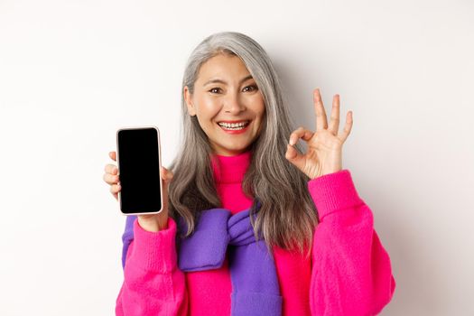 Online shopping. Fashionable asian senior woman showing blank smartphone screen and OK sign, smiling pleased, recommending app, white background.