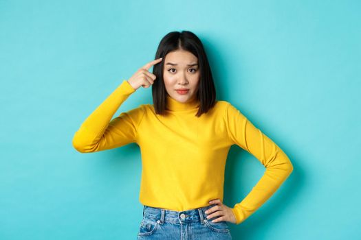 Are you stupid. Portrait of skeptical asian woman scolding someone crazy or dumb, pointing finger at head and staring at camera with dismay, blue background.