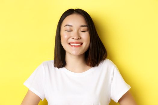 Close up of happy, romantic asian girl dreaming of something, close eyes and smiling delighted, standing over yellow background.