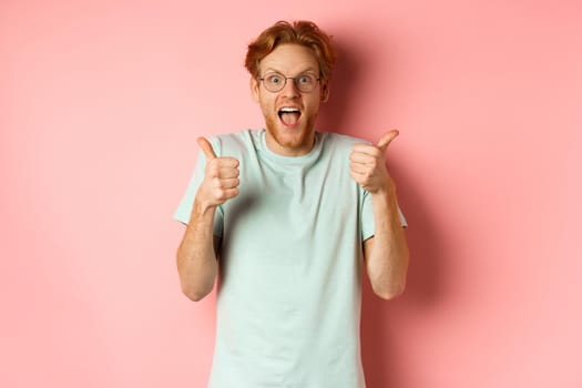 Amazed redhead guy looking impressed and excited, showing thumbs up in approval, praise awesome promo offer, standing over pink background.