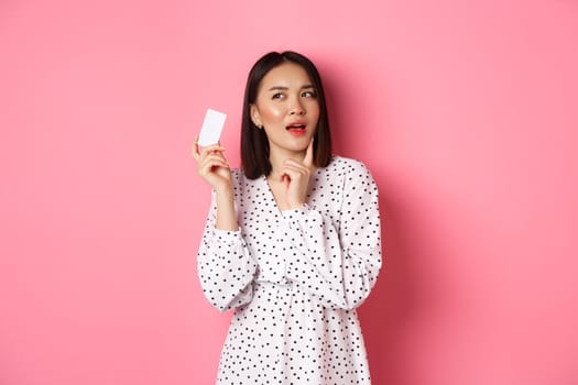 Shopping concept. Attractive asian woman thinking about buying presents, holding credit card and pondering, standing in dress against pink background.