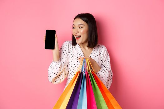Attractive asian woman showing smartphone app and shopping bags, buying online via application, standing over pink background.