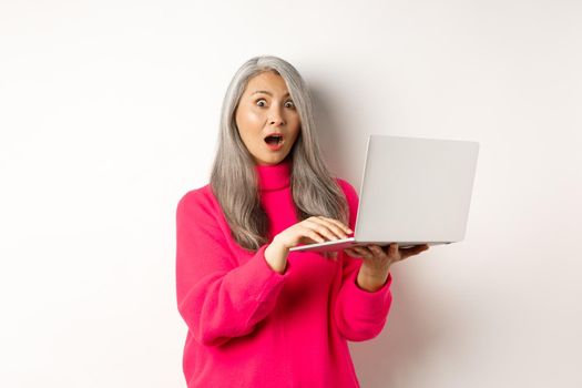 Surprised and impressed asian grandmother staring shocked at camera after reading news on laptop screen, standing over white background.