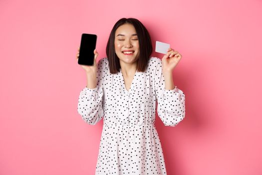 Joyful korean woman showing smartphone screen and credit card, paying for internet order, demonstrating online shopping app, standing over pink background.