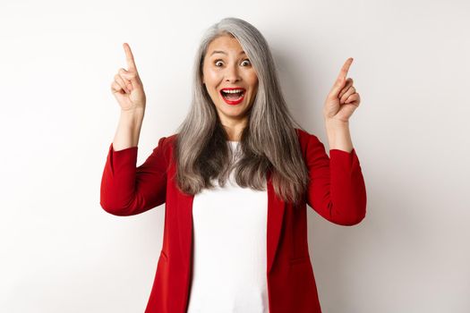 Mature asian businesswoman with grey hair, wearing red blazer and pointing fingers up, smiling surprised, showing promo offer, white background.